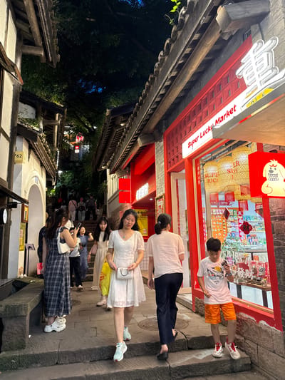 The image captures a bustling street scene in the Nan'an District of Chongqing, China, characterized by traditional Chinese architecture with overhanging wooden roofs and red accents. A group of people are present, with one person wearing a white dress and another in a blue plaid dress. Several individuals are holding smartphones and seemingly taking pictures. A shop with a sign reading "Super Lucky Market" is visible, featuring displays of merchandise and traditional Chinese lanterns. Other shops with red signage and decorations also line the street. The lighting suggests it is evening, with some areas illuminated by artificial lights and others shrouded in darkness.