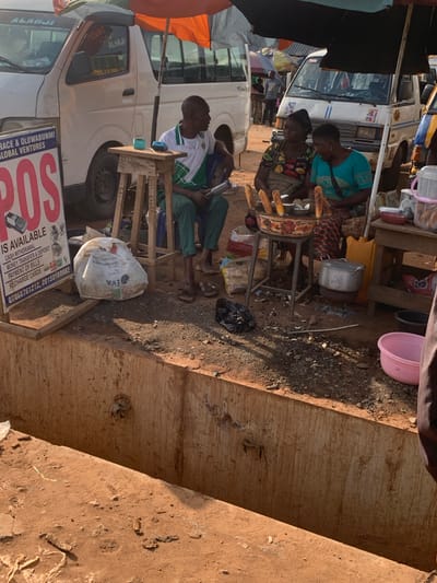 The image captures a street scene in Shagamu, Nigeria, likely in the late afternoon given the long shadows. The setting appears to be a bustling roadside market or transport hub, with several vehicles and stalls visible.
The primary subjects are several individuals, most of whom are women, engaged in selling goods. One woman in a blue top is focused on preparing food, possibly corn, which is displayed on a rack. Another woman in a patterned dress is also tending to the food stall. In the foreground, two men are seated on a wooden structure. One man, wearing a white and green shirt, is looking down, perhaps at a phone. The other man, in green trousers, is also seated.
Objects of note include a metal pot on a stand, various food items (likely corn), plastic containers, a black plastic bag, and a large white bag labeled "EVA". A white bus is parked prominently in the background, with its windows and doors visible. Another vehicle, partially obscured, is also present. The ground is a mixture of dirt and debris, characteristic of an outdoor market.
The activity taking place is commerce, with people selling and possibly buying goods. The scene conveys a sense of everyday life and economic activity in a Nigerian urban setting. The lighting suggests it is a sunny day, with the sun casting distinct shadows.
Visible text includes "EVA" on the white bag. There is also text on the side of the bus, partially obscured, but some words like "ALHAJI" and "YOUNG" are discernible. A large sign in the lower part of the image contains the word "SOS" in prominent pink lettering, along with smaller text that seems to advertise services like "RECHARGE CARDS", "PAYMENT OF BILLS", "MONEY TRANSFER & DEPOSIT", and "CASH WITHOUT DRAWER". A phone number "07068767212, 0750060082" is also visible. The overall impression is one of lively, informal commerce.