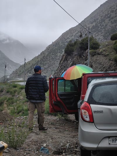 A man wearing a blue baseball cap, a dark puffer jacket, and tan cargo pants stands next to a red vehicle with its doors open, facing away from the camera. A rainbow-colored umbrella is propped on the roof of the red vehicle. A silver Maruti Suzuki car is parked behind the red vehicle. The scene takes place on a dirt road with sparse vegetation and rocky terrain, in front of a steep, mountainous landscape under a cloudy, overcast sky, consistent with the region of Darcha, India, known for its dramatic mountain passes.
