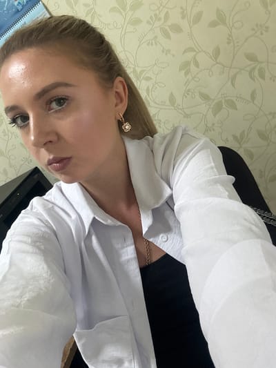 A close-up, selfie-style photo shows a young woman with blonde hair and light-colored eyes. She is wearing a white, collared shirt, unbuttoned, over a black top. A delicate gold necklace with a rose gold pendant is visible. She has a flower-shaped earring in her right ear, with a small pearl in the center and diamond-like accents. Her makeup includes mascara and lip gloss. The background features light green wallpaper with a subtle floral pattern. To the left of the woman, a black object, possibly a computer monitor or desk accessory, is partially visible. In the upper left corner, a portion of a calendar with Cyrillic script and the word "JUNE" is visible. The lighting suggests an indoor setting, possibly an office or a room in a residential building in Makiivka, Ukraine.
