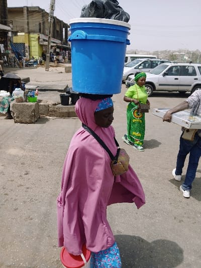 The image depicts an urban street scene in Masvingo, Zimbabwe, under bright, direct sunlight. In the foreground, a woman walks towards the viewer's left, balancing a large, bright blue plastic bucket with a white rim on her head. The bucket contains a black plastic bag. She wears a pink head covering over a blue patterned fabric, a long, loose pink outer garment, and a dark cross-body strap supporting a small, textured brown and gold bag at her right hip. Her left hand carries a white plastic container with a red lid.

To the right of the central figure, a second woman walks, wearing a lime green short-sleeved top, a green patterned skirt, and a green headscarf. She carries a small, light brown patterned purse. Further to the right, a third individual's arm and lower body are visible, holding a white rectangular tray that displays small, shiny objects. This person wears blue jeans and white sneakers.

In the midground, to the far left, a person is seated under a black umbrella, adjacent to concrete blocks used to display various items. The background features a paved, somewhat dusty street with several parked or moving white vehicles, including SUVs and sedans. Beyond the street, multi-story buildings with visible commercial storefronts line the road, some appearing weathered with exposed concrete and yellow paint. The sky is clear, and strong shadows are cast on the ground.