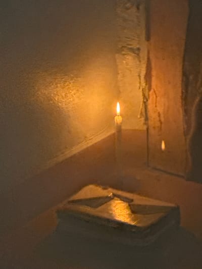 The image depicts a close-up, dimly lit scene with a focus on a small, bright flame from a candle. The flame is positioned horizontally and appears to be emanating from a wick. The surrounding area is textured and illuminated by the warm glow of the flame, creating a soft, diffused light. To the left of the flame, there is a textured surface that appears to be concrete or plaster, showing signs of wear and possibly dampness. Further back, a horizontal, lighter-colored surface is visible, also with a rough texture and hints of light reflection.
To the right of the flame, a distinct architectural element is present. It is a light-colored, angular structure with visible facets or a patterned surface. The texture here is granular, resembling rough stone or a textured wall finish. This structure is partially obscured by shadow. The overall lighting suggests it is either nighttime or a very dimly lit indoor space, with the candle being the primary light source. The textures and the quality of light evoke a sense of intimacy or perhaps a solemn moment. Given the location context of Badagry, Nigeria, this scene could be set in a historical or traditional dwelling, or a place of worship. No people are visible, and no text is present in the image. The activity is simply the burning of a candle, casting its light upon its immediate surroundings.