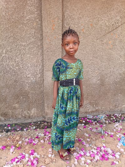 A young girl stands facing the camera in Gwagwarwa, Nigeria. She wears a floor-length, green and blue patterned dress with a black belt. The background is a rough, textured wall with a vertical seam. The ground is covered in red dirt and scattered with pink and white flower petals. There is trash visible near the base of the wall. The girl is wearing red sandals and has her hair braided.