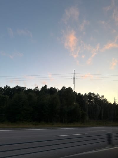 The image shows a view of a highway with a forest line on the right. A tall electrical pylon stands prominently in the distance above the trees. The sky is a pale blue with wisps of pink clouds, suggesting it is either early morning or late evening. The highway itself is paved and empty, with visible lane markings. A metal guardrail runs along the bottom of the image. The overall scene conveys a sense of travel or transit through a natural landscape. It is likely a rural or semi-rural setting in Rabbalshede, Sweden, given the presence of the highway and the dense forest.