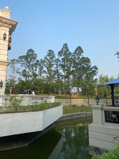 This media file depicts an outdoor scene with a calm atmosphere, likely in the late afternoon or early evening given the soft lighting and clear sky. The setting appears to be a well-maintained public space or park, possibly near a body of water, as a dark, calm pool of water is visible on the right side of the frame.
Several individuals are present, interacting with the environment. Two people are seen sitting on steps, engaged in conversation, while another person stands nearby, looking out towards the trees. Their postures suggest relaxation and casual interaction.
The scene features lush green trees with tall trunks and full canopies, providing a sense of natural beauty and shade. In the foreground, there is a modern architectural element, possibly a building or a decorative structure, made of light-colored stone. This structure has decorative lamps and a layered design. A walkway made of paving stones runs alongside the pool, bordered by neatly trimmed grass and small bushes. A striped awning, likely part of a nearby structure, is visible in the background.
The location cues suggest East Jakarta, Indonesia. The overall mood is peaceful and serene, implying a pleasant day for leisure and socializing in an urban green space. There is no discernible text within the image.