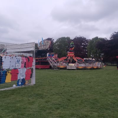 The image shows a fairground attraction on a grassy area, likely a park or common. In the foreground is a football goal with football shirts hanging from it for sale. The fairground ride has a large figure of a frog wearing a top hat on top of a red and gold carousel-like platform. There are steps leading up to a raised platform with a sign on it. The background consists of trees and an overcast sky. Based on the known location, the photo appears to be taken in Coventry, United Kingdom.