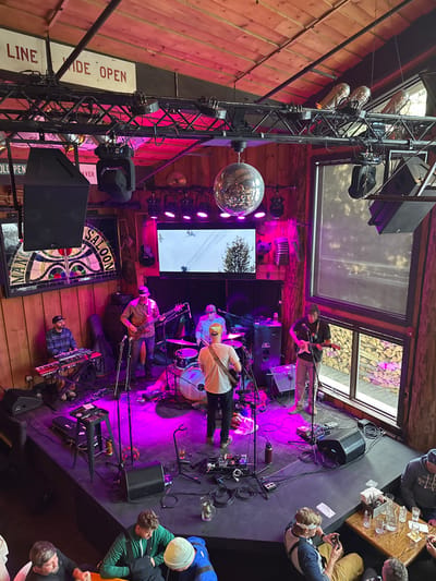 A band performs on a stage bathed in purple light. A drummer, guitarist, keyboardist, and other musicians are visible, surrounded by microphones and equipment. Above, a disco ball hangs near a stage light tower. A large TV screen displays a wintry outdoor scene. Patrons observe the show from the sides of the stage. Signs and decorative elements, like a stained-glass saloon sign, adorn the rustic wooden walls.