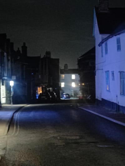 The image shows a street scene at night in what appears to be a historical area within East Suffolk, United Kingdom. The street is dark and wet, reflecting light from street lamps and building interiors. The buildings are a mix of architectural styles, including a white building with visible windows on the right, and darker, older structures on the left and further down the street. In the distance, lights shine through an opening between buildings. Vehicles are parked along the street, obscured by the darkness. A street lamp on the left illuminates the immediate foreground. No people are visible. The overall environment is urban and dimly lit.