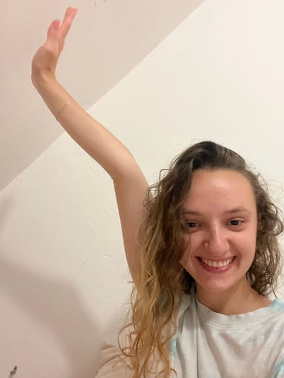 A young woman with light skin, light brown curly hair, and a bright smile is seen in a selfie. She is wearing a light-colored shirt. Her arm is extended towards the lower left corner of the frame, with a delicate gold bracelet visible on her wrist. The background is a plain white wall with subtle shadows, suggesting indoor lighting. There are no details to indicate this image was taken in Saarbrücken, Germany.