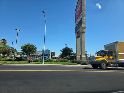 The image captures a commercial area in West Sacramento, United States of America, under a clear blue sky. A large, multi-columned sign for "GROCERY OUTLET BARGAIN MARKET" and "DELI TACO 24 HOUR DRIVE-THRU" is prominently displayed on the right side of the frame, rising high above the landscape. In the foreground, a paved road with double yellow lines runs horizontally across the bottom. To the right of the road, a bright yellow box truck with a silver trailer and an American flag decal on its rear is driving away from the viewer. The truck has the logo "ECF" visible on its side.
Behind the truck and across a grassy median, a parking lot is visible with several vehicles, including white sedans and a red sports car. A large white box truck is parked near a building. A fire hydrant, painted orange and red, stands near the grassy area. Further back, more trees and buildings, possibly storefronts, are seen. Several streetlights are scattered throughout the scene. A few individuals are visible in the distance, near the parking lot, seemingly interacting in the commercial environment. Lush green trees and bushes dot the landscape, interspersed with some flowering plants. The overall impression is a sunny day in a bustling commercial district.