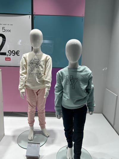 This image captures two child mannequins, positioned upright but rotated 90 degrees counter-clockwise, showcasing children's clothing within what appears to be a retail store display or window.
The mannequin positioned higher (or to the left in the rotated view) is dressed in a light mint green long-sleeved sweatshirt with the text "NY CITY" printed in black outline lettering. It is paired with dark blue or black trousers.
The mannequin positioned lower (or to the right in the rotated view) wears a cream-colored long-sleeved top featuring a black outline drawing of flowers with the words "love flowers" integrated into the design. This top is combined with light pink sweatpants.
Both mannequins have smooth, white, featureless heads and bodies, typical of retail displays. They stand on circular glass bases. The background behind the mannequins is composed of large rectangular panels in contrasting pastel colors: pink on the left and a muted blue on the right. Reflections are visible across the surface, suggesting a glass barrier like a shop window.
Visible text on a sign partially obscured by the lower mannequin includes "dès/vanaf/ab/από" (meaning "from" in several languages, including Greek) followed by "5,99€". Another price, "2,99€", is prominently displayed, along with the brand "Club ORCHESTRA". Near the feet of the lower mannequin, a small tag shows Greek text "ΦΟΥΤΕΡ" (sweatshirt) and "ΦΟΡΜΑ" (tracksuit/sweatpants), along with the number "2". The texts "NY CITY" and "love flowers" are visible on the respective garments.
The scene depicts a typical retail display, likely in Koridallos, Greece, given the Greek text. The time of day and weather are not discernible. No emotions are conveyed as the subjects are inanimate mannequins.