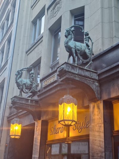 This is a medium-angle, eye-level, exterior shot of the historical building at Thomaskirchhof 12 in Leipzig, Germany. The facade of the building is made of light-colored stone blocks and features classical architectural elements.
Two ornate bronze sculptures of a goat being ridden by a cherubic child are mounted on cantilevered brackets projecting from the building. The sculptures are facing left and are positioned above arched entrances. The left sculpture appears to be slightly less detailed than the right one, but both depict the same subject.
Below the sculptures, on either side of the central entrance, are two large, hexagonal lanterns with yellow, illuminated glass panes. These lanterns hang from decorative metal brackets. The entrance itself has a sign that reads "Shero Style" in ornate, cursive lettering against a yellow background. Below this sign, a storefront window displays text, some of which is mirrored, suggesting it might be an "Asia Market" or similar establishment, with additional text visible such as "FOOD TO GO".
The building's upper floors are visible, showing rows of windows with light-colored frames. The architectural style is reminiscent of early 20th-century European design, with decorative molding and stonework. The overall impression is one of historical charm and artistic detail, characteristic of a well-preserved urban environment.