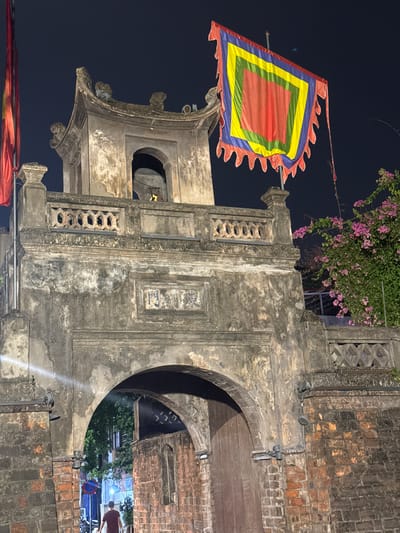 The image shows the imposing gate of the Temple of Literature (Văn Miếu-Quốc Tử Giám) in Hanoi, Vietnam at night. The gate is constructed of weathered stone and brick, with a traditional tiled roof and ornate carvings. A large, multi-colored flag, featuring concentric squares in red, yellow, green, and blue with a serrated edge, is prominently displayed on a pole to the right of the gate. To the right of the gate, a vibrant bougainvillea plant with pink flowers spills over a stone railing. The archway of the gate leads to a dimly lit inner courtyard, where a single individual in a maroon shirt is walking away from the camera. The sky is dark, indicating nighttime. The overall impression is one of historical grandeur and cultural significance.