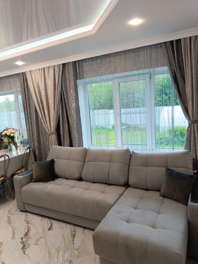 The photograph depicts an interior scene, likely a living room, in Andreyevo, Russia. The focal point is a large, light-gray sectional sofa with dark-gray throw pillows. Behind the sofa is a set of windows covered with sheer, vertically-striped curtains and heavier, gray-toned drapes. Outside the windows is a view of a green, wooded area and a white fence. To the left of the sofa, a dining table is partially visible, set with flowers and dining ware. The floor is covered in marble-patterned tiles. The ceiling is white with recessed lighting and a decorative molding. No people are visible in the frame.