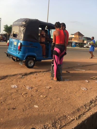 The image depicts a scene in Abakaliki, Nigeria. A blue "keke" or "tricycle taxi" is parked on a dirt shoulder beside a paved road. Two women stand beside the keke. The first woman is wearing a red top and pink and black patterned leggings and standing on a black plastic bag. The second woman behind her is also wearing a red top. Inside the keke are passengers, their faces partially visible. In the background across the road, another woman in blue clothing carries something wrapped in fabric. A building and other vehicles are visible in the distance. The ground around the keke is dirt with scattered debris.