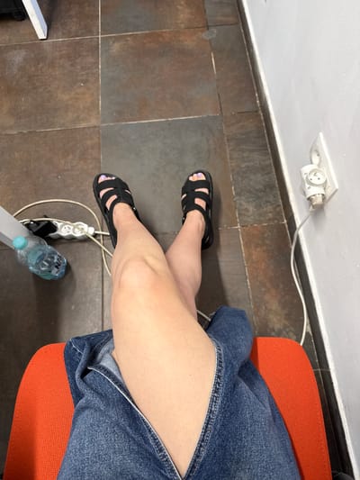The image shows a person's legs and feet from a top-down perspective, suggesting they are sitting or standing. They are wearing black sandals with a minimalist design and a denim skirt or shorts. The floor is tiled with dark brown square tiles. A white electrical extension cord with multiple outlets is visible on the floor, with one plug inserted into a wall socket. A plastic water bottle with a blue cap rests near the extension cord. The scene appears to be indoors, possibly in an office or home, given the presence of electrical outlets and furniture (an orange seat is partially visible). The lighting suggests it is daytime. There is no discernible weather information or strong emotional cues. The location context is Łódź, Poland, but there are no specific visual cues to confirm this.