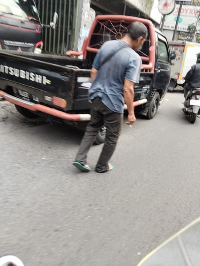 The image shows a man in a blue t-shirt and dark pants walking on a street in Cimahi, Indonesia. He is wearing green flip-flops. Behind him, a black pickup truck with a red roll cage is parked. The truck has "MITSUBISHI" written on the side. A white truck is visible in the background, as is a person riding a motorcycle. The scene appears to be a busy street with traffic. The overall lighting suggests it is daytime, possibly overcast.