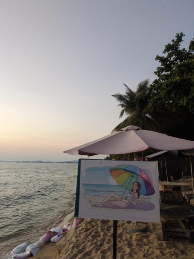 The scene depicts a beach in Phú Quốc, Vietnam. A signpost featuring an illustration of a woman with an umbrella is visible. Sandbags are placed near the shoreline to prevent erosion. The sea stretches out into the distance. A tree is visible in the background. An umbrella partially obscures the sign. The sky is pale and clear.