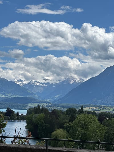 A scenic view of the city of Thun, Switzerland, is presented, featuring a vast expanse of the sky adorned with fluffy white clouds against a brilliant blue backdrop. Below, majestic snow-capped mountains rise, their peaks partially obscured by dramatic cloud formations. The midground is a tapestry of lush green trees and rolling hills, interspersed with the tranquil waters of a lake. Small boats dot the lake's surface, and houses are nestled amongst the foliage, hinting at the vibrant life within the city. In the foreground, a stone wall and railing offer a vantage point for this breathtaking panorama.