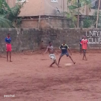 Four people are playing soccer on a dirt field. In the background is a weathered wall with the words "UNITY CLUB" painted on it. There are buildings and trees visible behind the wall. The photograph includes the name "paschal" in the lower left corner. Based on this information, the image is likely taken near a local Unity Club playing field. The location is possibly Ukwu-Abwa, Nigeria, as stated.