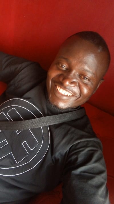 A smiling Black man in a black shirt with a white outlined graphic sits against a red backdrop. A black strap crosses his chest. The image is taken at an upward angle, showcasing his cheerful expression and attire. The focus is primarily on the man's face, highlighting his smile and the details of his features.