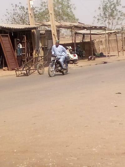The image depicts a daytime street scene in Tudun Murtala, Nigeria. A male individual, dressed in a light blue traditional garment and a green cap, operates a dark-colored motorcycle moving from right to left across an asphalt road in the foreground. The road transitions to a dusty, unpaved shoulder on both sides.
On the left side of the road, a makeshift commercial stall with a corrugated metal roof stands open-fronted. Inside, a second male wearing a light blue and dark brown shirt is visible. Adjacent to the stall, a two-wheeled metal cart or trailer is positioned. Various metal objects, including what appears to be door frames, are stacked within and leaning against the stall structure. A long, brown, inclined object, possibly a ramp or large plank, rests against the stall's front.
On the right side of the road, further down the shoulder, another similar open-fronted shelter with a corrugated roof is present. A white scooter is parked on the dusty ground near this shelter, alongside other indistinct dark objects. A low, light-colored wall runs parallel to the road. The background reveals additional low-rise structures and sparse, light green foliage against a hazy sky. The overall environment appears dry and dusty.