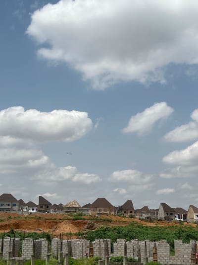 An aerial view captured from a distance shows a residential development under construction in Kubwa, Nigeria, beneath a partially cloudy sky. The foreground is dominated by the skeletal framework of unfinished buildings, characterized by concrete columns and cinder block walls. Beyond this construction zone, a landscaped hillside rises, dotted with completed and near-complete houses featuring tiled roofs and modern architectural designs. These homes are situated on tiered levels, with some appearing to be two-story structures. The landscape is a mixture of exposed reddish-brown earth and patches of green vegetation. In the sky, a commercial airplane is visible in the distance, appearing as a small, dark silhouette against the lighter clouds. The overall impression is one of ongoing urban expansion and development within a suburban setting.