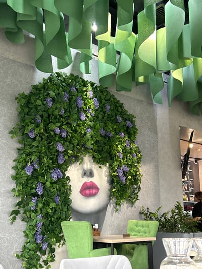 The image depicts the interior of a commercial establishment, likely a restaurant or cafe, located in Raduga, Russia. The central feature is a large, stylized female face mural on a textured light grey wall, with bright pink lips and artificial green foliage adorned with clusters of small purple flowers serving as the hair. The foliage cascades down the wall to the left of the face. Above, numerous vertical strips of light green material, curled and undulating, are suspended from the dark ceiling, creating a decorative canopy.

In the mid-ground, a seating area includes two bright green upholstered chairs with button tufting positioned around a light-colored table with a dark frame. A small "RESERVED" sign rests on this table. Partially visible in the bottom left is a white chair. To the right of the seating area, a grey planter holds several live green plants with dark leaves. Further in the background, a reflective surface shows distorted reflections of overhead lighting. To the far right, a partial view of a bar or shelving unit displays various bottles and glassware. A person with dark hair, wearing a dark top, is partially visible near these shelves, facing away from the camera. The immediate foreground on the right shows out-of-focus glass carafes or pitchers. The overall environment is modern and clean, with a prominent natural aesthetic.
