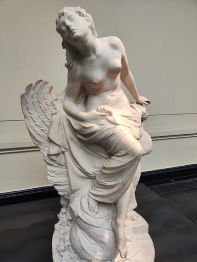 The image shows a marble sculpture of a semi-nude female figure. She is seated, draped with fabric that covers part of her body. She appears to be riding or supported by a winged creature, likely mythical in nature. The statue is located indoors, evidenced by the wall and the flooring in the background. It is at the Museo Nacional de Bellas Artes in the city of Santiago, Chile.