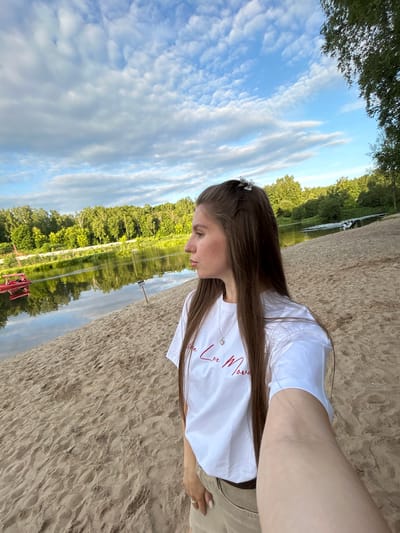 This media file captures a young woman on a sandy beach, with a serene lake and lush green trees forming the backdrop. The image is oriented sideways, with the sky and water occupying the left portion of the frame. The sky is a brilliant blue, dotted with wispy white clouds, suggesting a pleasant day. The water of the lake is calm, reflecting the surrounding trees and the sky. The trees are dense and green, creating a natural, tranquil environment.
The woman, with long, dark brown hair, is wearing a white t-shirt with red text on it, and light-colored shorts or pants. She is looking down and appears contemplative. A small, sparkly accessory is visible in her hair, and a necklace with a small pendant hangs from her neck. The sand is a light beige color, with visible footprints. In the distance, a red object, possibly part of a small boat or watercraft, is partially submerged in the water.
The lighting suggests it is either late afternoon or early morning, given the soft quality of the light and the angle of the sun. The overall mood is peaceful and relaxed, evoking a sense of quiet enjoyment of nature. The text on the woman's shirt is partially visible and appears to say "Love Moves." The location context provided indicates Klin, Russia.
