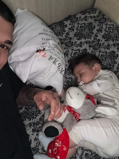 A top-down, close-up shot depicts a baby sleeping on a patterned blanket next to a plush grey donkey toy with a red bow and an embroidered red heart. The baby has a pacifier in its mouth. A white pillow with Bulgarian text is also present. Part of a person's face and arm are visible in the foreground. The text on the pillow includes the word "KЪПА", which could be translated as "bathe". There is no information available in the image to ascertain the location of the image, including whether it is in Krichim, Bulgaria.