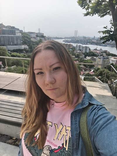 A woman with blonde hair is taking a selfie with a scenic background of Vladivostok, Russia. She is wearing a pink t-shirt with a graphic of an eagle and a denim jacket. A green backpack is visible. The background shows buildings, a body of water, and bridges. The setting appears to be an elevated vantage point overlooking the city.