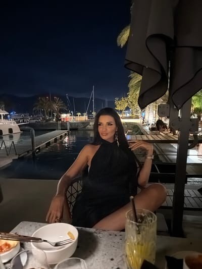 A woman in a black dress is seated at a table outdoors at night, with a marina in the background. The woman has long dark hair, is wearing makeup, and jewelry, including earrings and bracelets. She is looking to her left with her legs crossed. On the table in front of her are bowls of food, a fork with a piece of food, and a glass with a straw. The marina is lit with lights and features boats and palm trees. The location is Tivat, Montenegro.