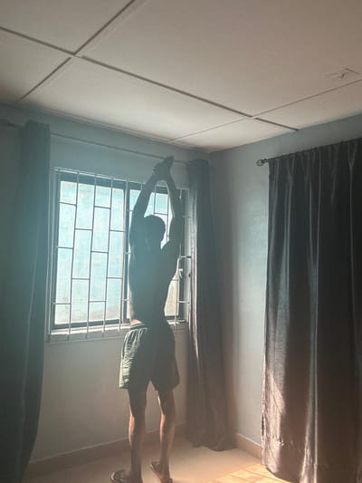 A person is standing in a room with their arms raised above their head, in front of a window. The window has metal bars across it. The room has dark curtains on either side of the window and a tiled ceiling. Sunlight is coming through the window, casting shadows on the floor. The person is wearing shorts. The scene appears to be located in Badagry, Nigeria.
