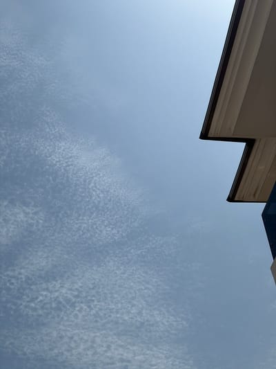 This is a low-angle, outdoor, daytime photograph taken in Igboefon, Nigeria, looking up at the sky. The sky is a pale blue with scattered, wispy white clouds that form a textured pattern across the left side of the frame. The right side of the image features the corner of a building's overhang. The overhang has multiple layers of trim, with the outermost layer appearing dark brown or black, and the inner layers a light, creamy color, possibly painted wood or a similar material. A dark blue vertical surface of the building is visible at the bottom right edge of the frame. There are no people or animals present in the image, and no discernible actions or interactions are taking place. The scene depicts an architectural element against a cloudy sky.