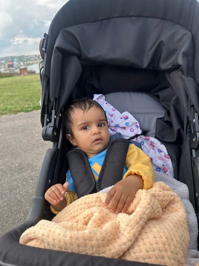 The image depicts an infant seated in a black baby stroller, located outdoors on a paved path in Great Lines Heritage Park, Chatham, United Kingdom. The child, appearing to be of South Asian ethnicity, has dark hair and brown eyes, wears a blue and yellow long-sleeved top, and is secured by black harness straps. A light orange textured blanket covers the infant's lower body, with a white patterned blanket visible behind. The stroller's canopy is partially extended, showing a "mybabiie" brand label. To the left, a grassy hill slopes upwards. The background reveals a panoramic view of the Medway Estuary, with urban buildings and distant hills under an overcast sky. The infant looks directly forward with a neutral expression.