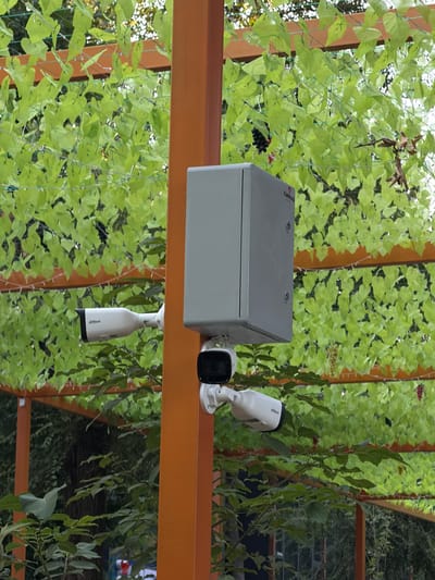 The image shows an outdoor scene with green foliage. A light grey electrical junction box is mounted on an orange beam. Two white security cameras are attached to the junction box. One camera is positioned to the right of the junction box, and another smaller black camera is attached below the first. The junction box has a label with the text "KANHAD" printed on it. The background consists of dense green leaves, possibly artificial, with some real branches interspersed. The lighting suggests it is daytime. The scene appears to be in an outdoor setting, possibly a garden or patio area, given the foliage and the mounting of the equipment. There are no people visible in the image.
