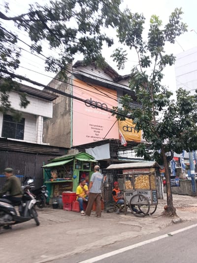 The scene depicts an urban street environment in Bandung City, Indonesia, under an overcast sky. An asphalt road occupies the foreground, bordered by a concrete sidewalk on the right. Numerous overhead power lines traverse the upper portion of the frame.
On the left, a two-story building with white tiled cladding is partially visible. Centrally located in the background is a multi-story building featuring a large, light-pink banner displaying "dema depo alat mebel" (dema furniture tools depot) and listing "blockboard, triplek, tipis, dan aksesoris mebel lain-lain" (provides: blockboard, plywood, thin, and other furniture accessories). A smaller yellow banner with similar text is below it. This building has a traditional sloped roof. To its right, a modern building is partially visible.
On the sidewalk, a green-painted vendor kiosk is positioned. Adjacent to it are stacked red plastic crates. To the right of the kiosk, a two-wheeled wooden cart, labeled "KRIPIK SINGKONG" (Cassava Chips) and "Aroma," displays bags of chips. A white tarp is strung above this vending area. A small tree with exposed roots stands next to the chip cart.
Four male individuals are present. On the far left, a person in a green top and cap operates a dark-colored scooter, moving leftward and exhibiting motion blur. Near the green kiosk, one male subject in a yellow t-shirt and blue jeans is seated on a red crate, facing right. Another male subject, wearing a light blue t-shirt and brown pants, stands with his back to the camera, appearing to engage the seated individual. Near the chip cart, a third male subject in an orange t-shirt and dark pants is seated on a low stool, facing left.