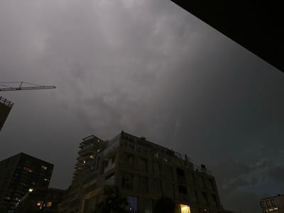 The image captures a dramatic cityscape at dusk or during a storm in Amsterdam, Netherlands. The sky is overcast and dark, with visible cloud formations. In the upper left portion of the frame, a construction crane extends horizontally across the sky. Below, several buildings are visible. To the left, a tall, dark, modern building with illuminated windows stands prominently. To the right and slightly forward, a multi-story brick building with a unique stepped facade and illuminated windows is featured. On the rooftop of the brick building, two figures can be seen, appearing to be standing and observing the surroundings. The scene conveys a sense of urban activity under moody weather conditions.