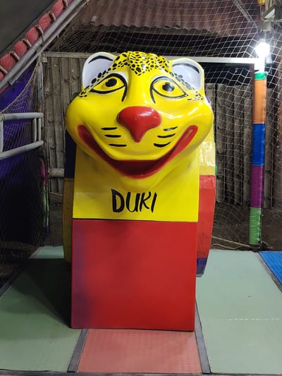 The image shows a brightly colored, cartoonish jaguar statue labeled "DURI." The statue is primarily yellow with black spots on the head, a red nose, and a red rectangular base. It is situated indoors on a multi-colored padded surface, likely within a play area. Background details include a metal fence, wooden planks, netting, and a pole wrapped in colorful tape. The location is likely a play area in Trujillo, Peru.