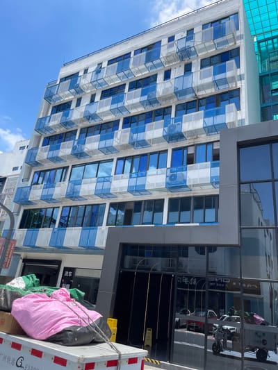 This image depicts a modern multi-story building in Panyu District, China, characterized by its white facade and alternating blue and white balcony enclosures. The building features numerous large windows, many of which reflect the blue sky and clouds. The lower levels of the building appear to house commercial establishments, with signs visible, including one that reads "洗车" (car wash).
In the foreground, a white flatbed truck is partially visible, loaded with various items covered in green and pink tarps. Notably, a large pink bundle is secured with ropes. The truck's side displays Chinese characters and the word "EXPRESS." The reflection in the building's large glass doors shows street-level activity, including vehicles and a three-wheeled scooter. The overall scene suggests a commercial area within the city, likely during daylight hours under a clear sky.