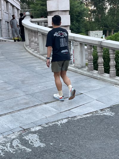A man walks away from the camera on a grey stone pathway. He is wearing a black t-shirt with white graphics, olive green shorts, cream-colored socks, and white and red sneakers. Other people are visible in the background, including a man in a grey t-shirt and grey pants standing on a raised platform to the left of the frame, and two individuals in dark clothing standing near a white stone railing. The environment appears to be outdoors, possibly in a park or public area, with trees and buildings in the distance. The pathway is bordered by a decorative stone railing with turned balusters. The ground is paved with large, rectangular grey stones.