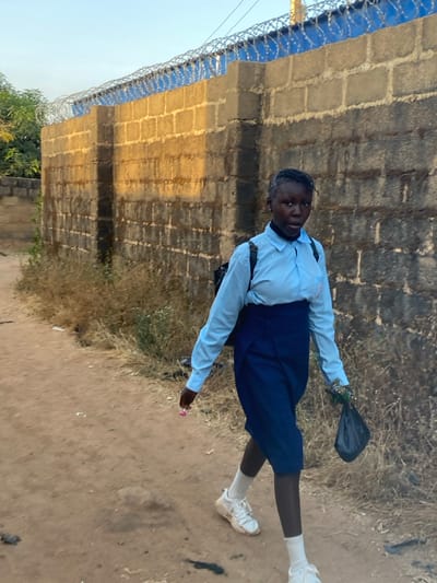 An individual walks along an unpaved dirt path in Kirana, Nigeria. The subject, a young person with dark skin and braided hair, wears a light blue long-sleeved collared shirt, a navy blue high-waisted skirt, white socks, and white athletic shoes. A dark backpack is visible on their back. The individual holds a pink lollipop in their left hand and a crumpled black plastic bag in their right hand. They are looking directly forward, towards the camera. To the right of the path stands a tall wall constructed from grey concrete blocks, partially covered with blue material and topped with razor wire. Sunlight illuminates the upper portion of the wall. Dry, sparse vegetation and some ground debris are present along the path and at the base of the wall. A small amount of green foliage is visible in the upper left background against a clear, light blue sky.