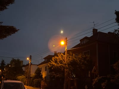 The image depicts a residential street scene in Poznan, Poland at dusk or night. The sky is a deep blue-grey, with a sliver of moon visible in the upper center. A street light with a warm yellow glow illuminates a section of the scene, creating a noticeable lens flare. In the foreground, the hood and windshield of a white car are visible on the left. Behind the car, a residential street runs towards the right.

Several houses are visible, characterized by tiled roofs and windows. Some houses are painted lighter colors, while one prominent house on the right is a dark reddish-brown. Trees and bushes with dense foliage are scattered throughout the scene, with some illuminated by the street light and house lights. Electrical wires crisscross the sky above the houses and trees. A traffic sign, yellow and triangular with a black "X" marking, is positioned on the right side of the frame, indicating a crossroads or intersection. The overall impression is a quiet, urban residential area at night.