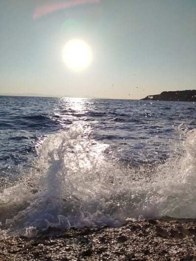 A vertical, eye-level shot captures a wave crashing onto a rocky shore near Karaburun, Turkey, under a bright, hazy sun. The sun, positioned in the upper left quadrant of the frame, is a brilliant, blown-out circle casting a strong glare and reflections across the choppy, dark blue ocean. The water is textured with small waves and scattered white foam. In the foreground, a large wave is breaking, sending white spray and droplets into the air. The shore is comprised of dark, rough rocks, some of which are wet from the spray. In the distance, a dark, tree-covered landmass is visible on the horizon. The sky is a pale, almost white blue, with a slight pinkish hue near the sun due to lens flare. The scene appears to be a natural coastal environment.