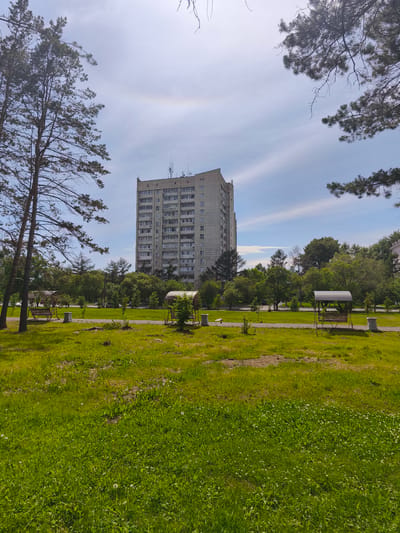 The image depicts an outdoor scene with a tall, light-colored apartment building in the background. The building has many windows and appears to be made of concrete panels. In front of the building, there is a park-like area with green grass and trees. There are two swings with canopies visible in the park. The sky is blue with some scattered clouds, and a faint rainbow is visible. The foreground is dominated by a grassy field with some scattered dandelions. The overall impression is a residential area in a city. Based on visual cues and the context provided, this scene is located in Komsomolsk-on-Amur, Russia.