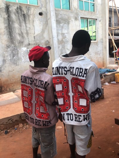 The image captures two young individuals, likely boys, from behind, standing outdoors in what appears to be a residential or developing area. The photo is taken in bright daylight.
The person on the left is wearing a white long-sleeved shirt with a collar, light grey shorts, and dark shoes or sandals. Their hair is short and dark.
The person on the right is wearing a light brown or tan long-sleeved shirt, distressed dark denim shorts, and a red baseball cap worn backward.
Both shirts feature a prominent design on the back. A large, stylized red number "88" with a black outline and white dots fills a significant portion of the back. To the left of the "88" (from the viewer's perspective), the text "SAINTS-GLOBAL" is printed vertically in black letters. To the right of the "88", the text "BORN TO BE WIN" is also printed vertically in black. Both shirts have similar patterns on the sleeves, extending from the main design.
The background consists of unpainted, raw concrete buildings, suggesting construction or unfinished structures. Several windows with light green or blue frames are visible in the building behind the individuals. In the upper left background, some bamboo scaffolding or construction materials can be seen. The ground is a reddish-brown dirt surface, with small stones and debris scattered around.