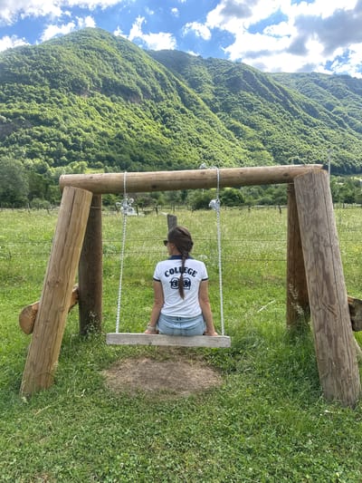 A young woman with dark hair in a braid, wearing a white t-shirt with the word "COLLEGE" printed on it in dark letters and denim shorts, sits facing away from the camera on a wooden swing. The swing is supported by a rustic wooden frame constructed from thick logs. The scene is set in a grassy field with scattered wildflowers. In the background, a dense, green, tree-covered mountain rises under a partly cloudy blue sky. The location is a rural area near Gusinje, Montenegro.