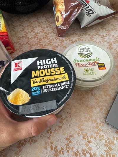 A close-up photograph depicts a human hand holding a black-labeled plastic container of "K CLASSIC HIGH PROTEIN MOUSSE Vanillegeschmack" (Vanilla flavor) with "20g PROTEIN PRO BECHER" and "FETTARM & OHNE ZUCKERZUSATZ*" indicated on the lid. A timestamp "03/04/26 S 11:16/9/07 123" is visible on the container's lid. To the right, on a light brown tablecloth with a white lace-like floral pattern, sits a clear plastic container of "Caña Nature Guacamole Mexicano" (96% AGUACATE) bearing a "SABOR DEL AÑO 2026" sticker. In the background, out of focus, are various packaged food items, including a brown package containing a pastry and other partially obscured packages. The silver edge of an electronic device is visible in the lower right corner. The scene is indoors in Ciorescu, Moldova.