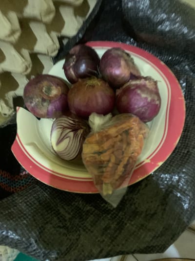 The image depicts a red and white plate filled with red onions and a clear plastic bag containing dried chili peppers. The plate is positioned on top of a dark, possibly textured cloth. To the left of the plate is a partially visible egg carton. The objects are situated in Keffi, Nigeria.
